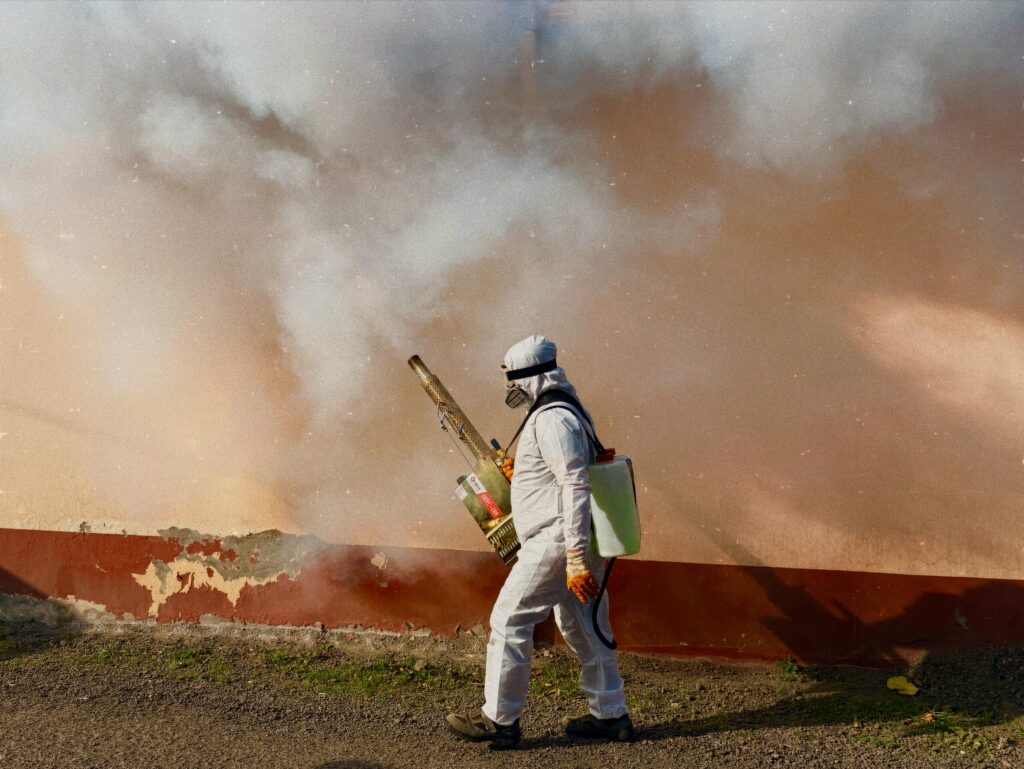 A worker in a full protective suit fumigates an area outdoors, spraying pesticide.
